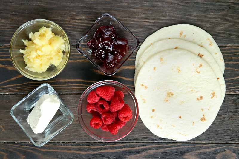 Overhead horizontal image of small glass dishes of cream cheese, crushed pineapple, jam, and whole raspberries, with a small stack of flour tortillas fanned out to the right, on a dark brown wood surface.