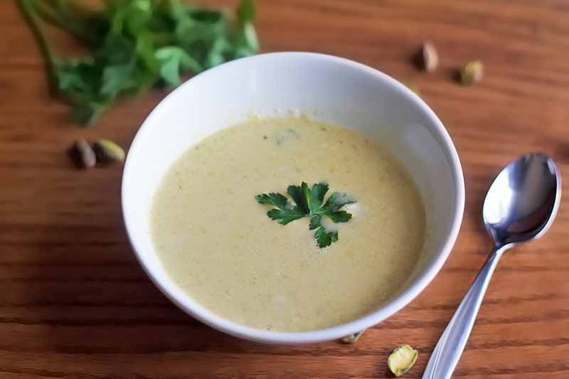 A white bowl of homemade cream of pistacho soup with a fresh herb garnish and a spoon to the right, with a sprig of flat-leaf parsley and scattered on nuts on a brown wood surface.