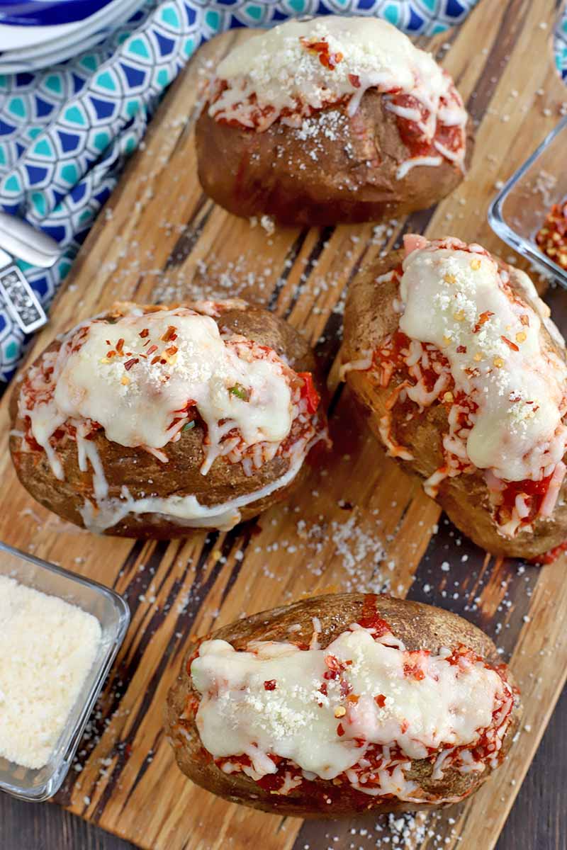 Four baked potatoes stuffed with marinara sauce and melted cheese, o a wooden serving board with a small square glass dish of grated Parmesan, on a light and dark blue cloth with a repeating triangular pattern, on a dark brown wood surface.