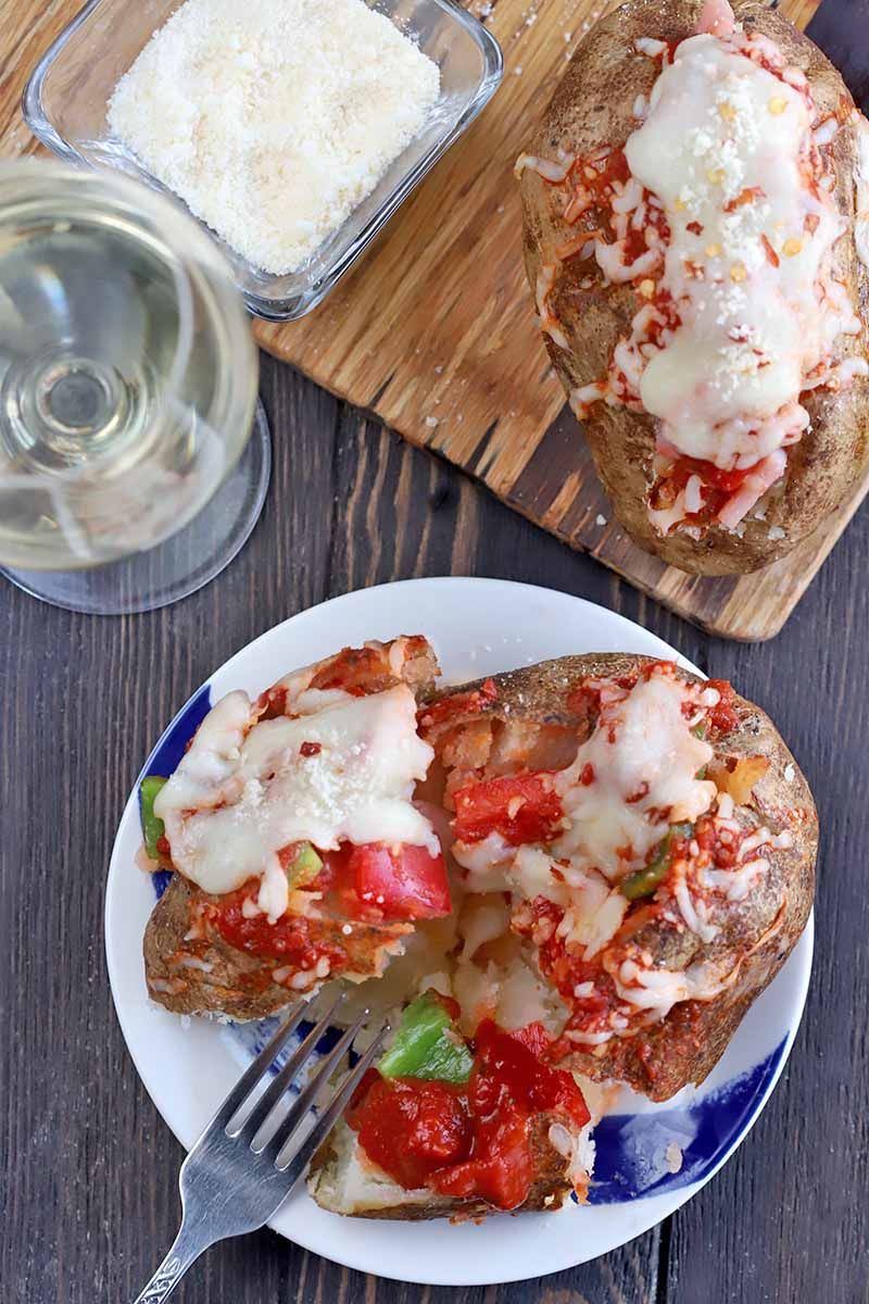 Overhead shot of two baked potatoes with pizza fillings, on broken open on a plate with a fork and another still intact on a wooden serving platter beside a small glass dish of grated Parmesan, with a glass of white wine, on a dark brown wood surface.