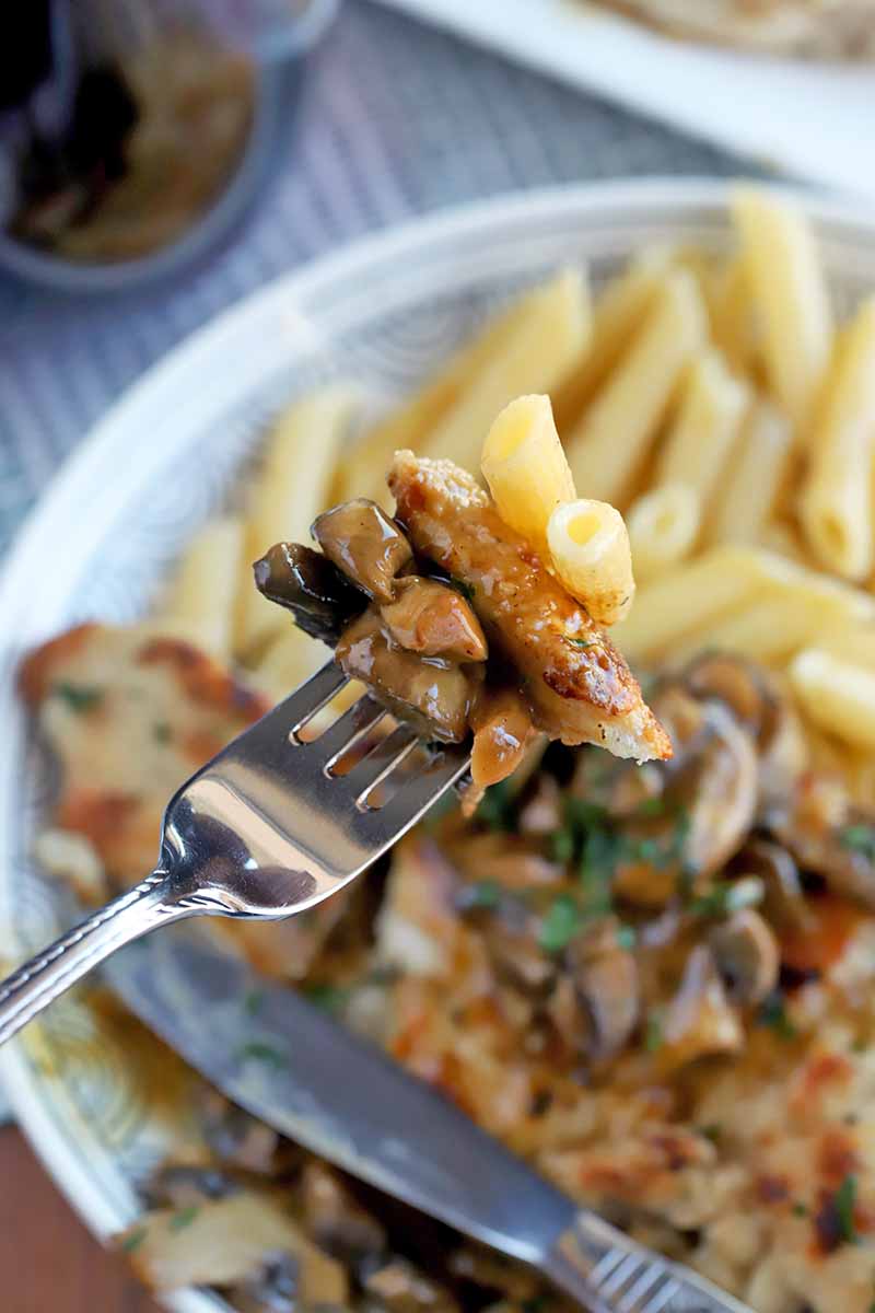 A forkful of chicken marsala and penne pasta is being held up to the camera, with more on a plate in shallow focus in the background, on a blue and white cloth with a stemless glass of red wine and a knife.