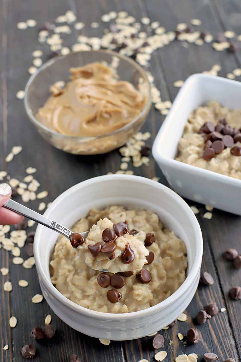 A hand with a manicured silver fingernail holds a spoonful of oatmeal topped with chocolate chips, with two white bowls of the hot breakfast cereal and a glass bowl of peanut butter in the background, on a dark brown wood surface with scattered uncooked oats and dark chocolate.