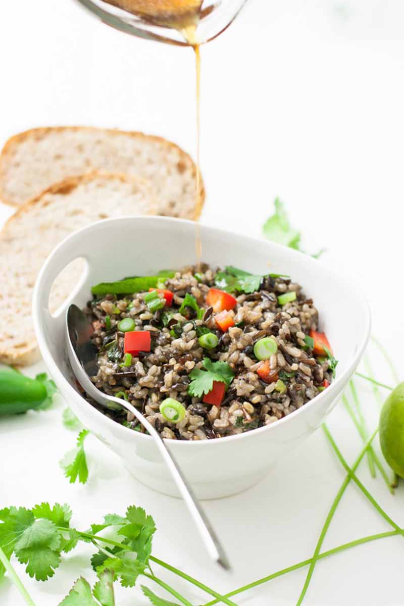 Vertical image of a grain salad in a white bowl with a spoon with dressing being poured into it, with bread and fresh ingredients on a white surface.
