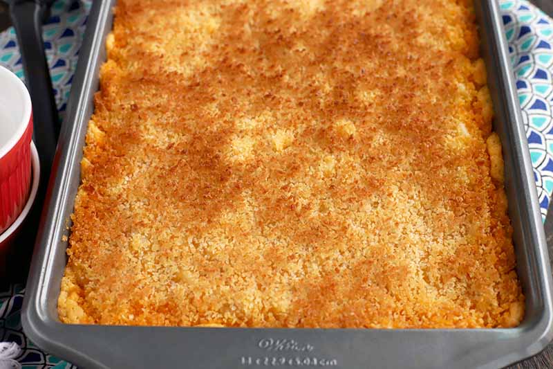 Baked macaroni with a toasted breadcrumb topping in a rectangular metal baking pan, on a dark and light blue patterned cloth surface with two red and white ceramic ramekins to the left of the frame.