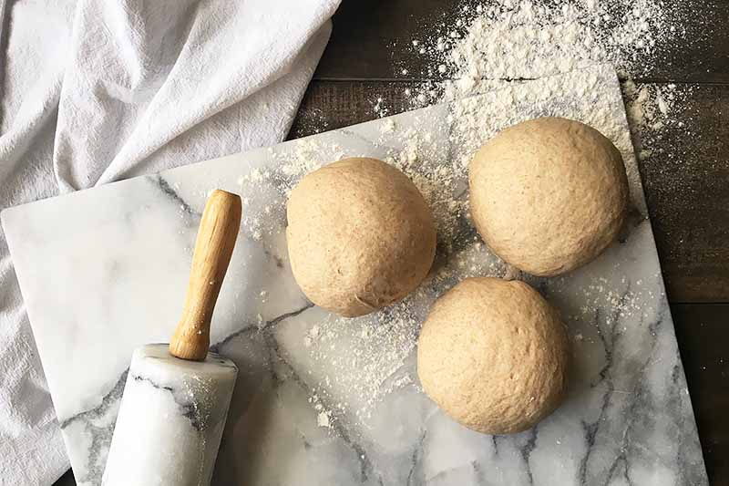 Horizontal image of bread dough rounds on a marble surface with a marble rolling pin.