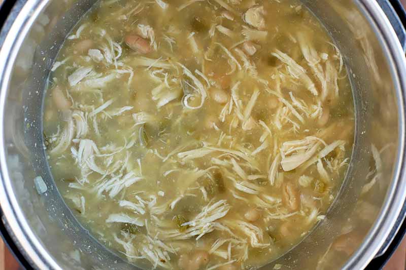 Closely cropped overhead shot of shredded chicken with cannellini beans and cooked peppers in a greenish broth fills the metal insert of a slow cooker, on a wood surface.