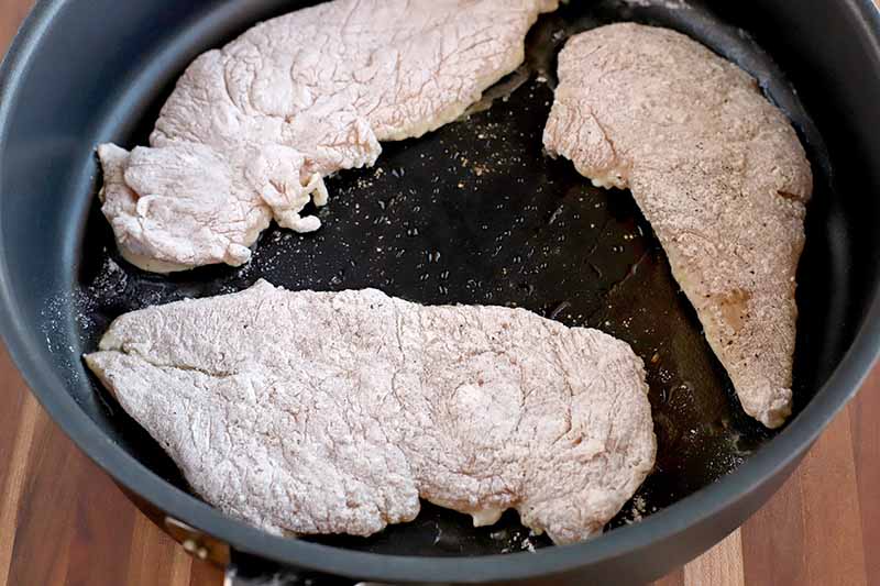 Chicken breasts dredged in flour, cooking in a nonstick frying pan, on a brown wood surface.