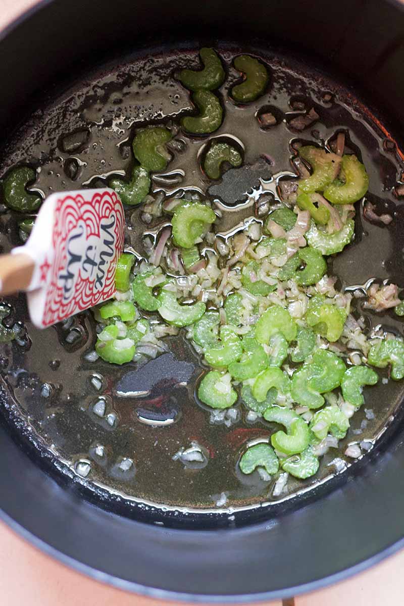 Vertical closely cropped overhead shot of a nonstick cooking pot with oil, chopped celery, and minced garlic at the bottom, being stirred with a silicone spatula that says "Mix It Up."