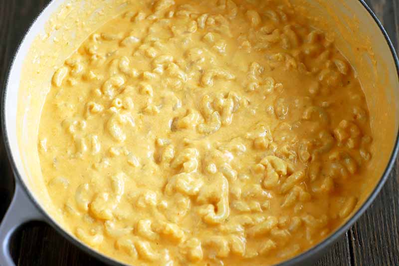 Cooked elbow macaroni coated in a thick cheese mixture in an enameled pot, on a dark brown wood surface.