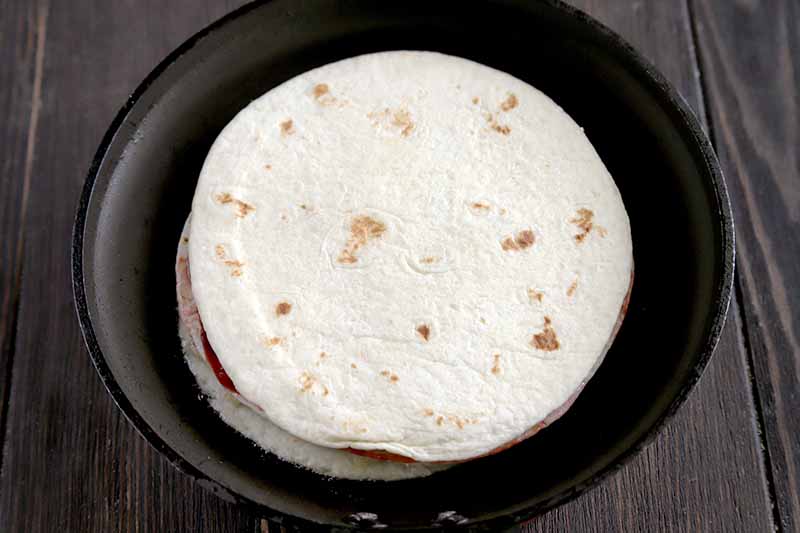 Horizontal overhead image of two flour tortillas with a sweet fruit and cream cheese filling in between, in a small nonstick frying pan on a brown wood surface.