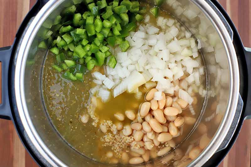 Overhead shot of a black plastic slow cooker with a stainless steel insert, filled with cannellini beans, chopped onion and green bell pepper, salsa verde, green hot sauce, chicken broth, and seasonings, on a striped wood surface.