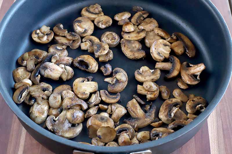 Sauteed sliced crimini mushrooms in a large nonstick frying pan, on a brown striped wood surface.