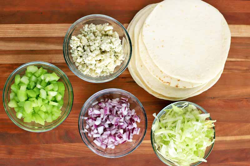 Overhead shot of four small glass bowls of chopped celery, crumbled blue cheese, chopped red onion, and shredded lettuce, with a small stack of flour tortillas, on a brown and beige wood surface.