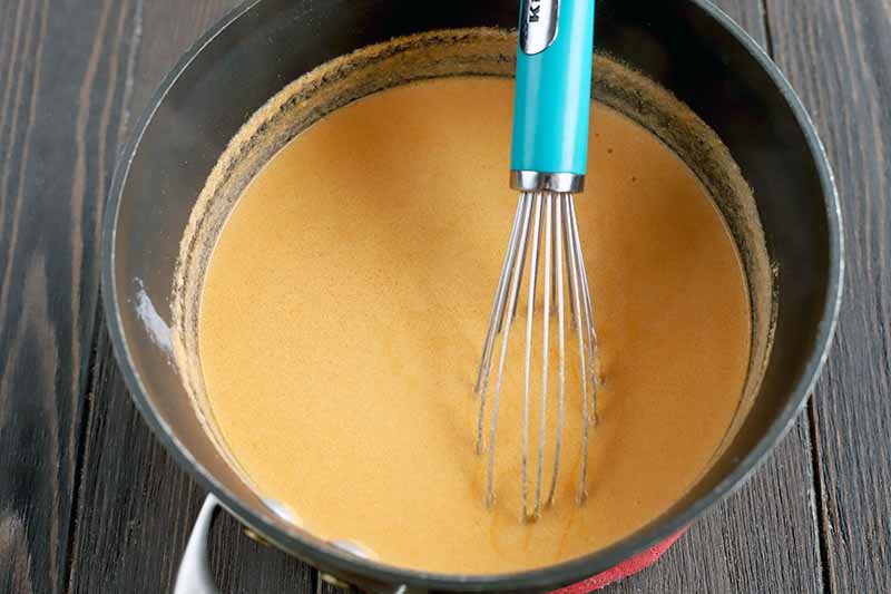 A wire whisk with a light blue handle is being used to stir a melted cheese mixture in a nonstick saucepan, on a dark brown wood surface.