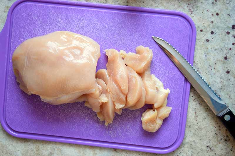A boneless, skinless chicken bread is being sliced o a purple plastic cutting board with a serrated knife with a black handle, on a speckled beige granite countertop.