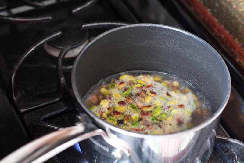 A stainless steel saucepan of pistachios is boiling on a gas stove, in frothy water.