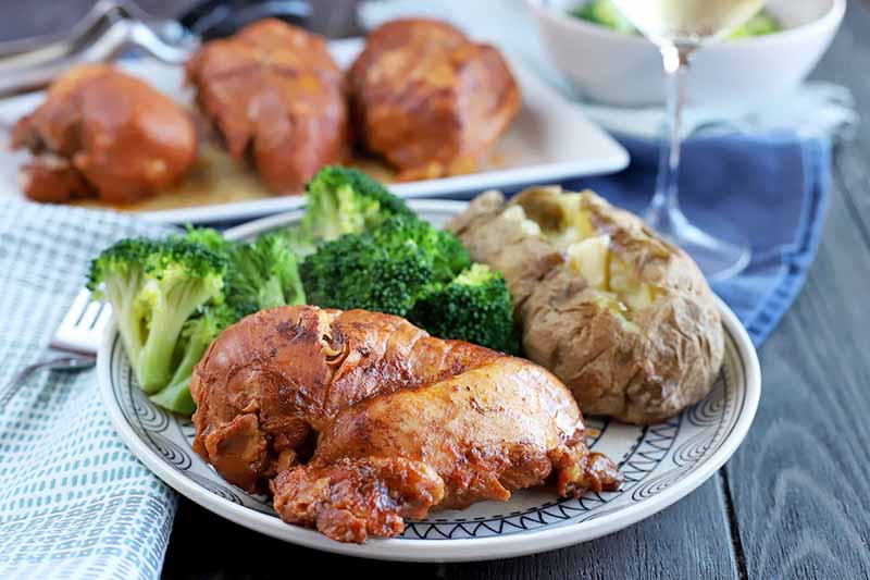 A white plate with a black decorative pattern is topped with barbecue chicken breast, a baked potato, and steamed broccoli, with a folded blue and white cloth napkin and silverware to the left, and a serving platter of chicken beside a bowl of vegetables with a glass of white wine in soft focus in the background, on a blue cloth on top of a dark brown wood table.