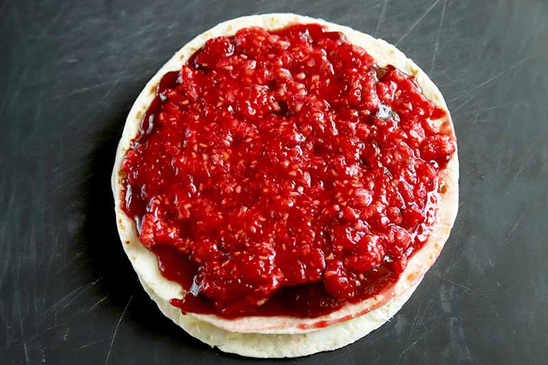 Overhead horizontal image of a stack of flour tortillas with filling in between and a layer of mashed fresh raspberries on top, on a scratched gray surface.