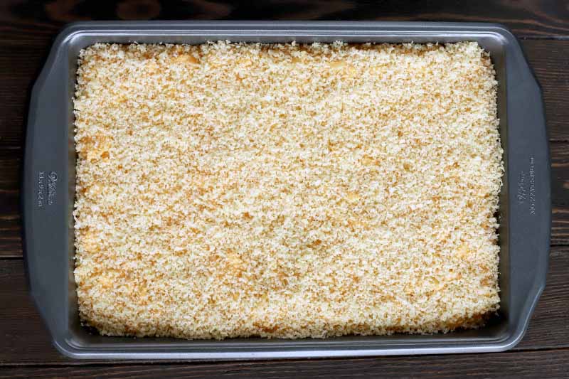 Overhead shot of a metal baking pan filled with pasta with panko bread crumbs evenly distributed on top, on a dark brown wood surface.