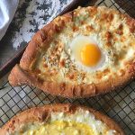 Horizontal image of baked bread with browned cheese and eggs next to a countertop with flour.