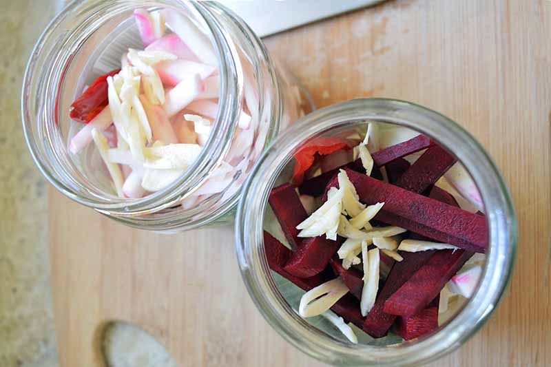 Overhead image of two glass jars filled with sliced turnips and beets, with some sliced garlic on top, on a wooden cutting board on a beige countertop.