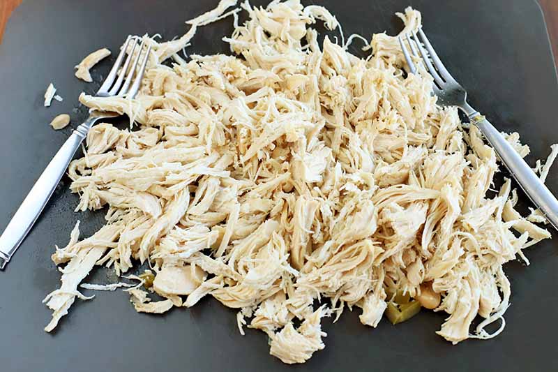 Shredded cooked chicken breast with two forks and a few stray pieces of cooked green bell pepper and onion, on a black plastic cutting board.