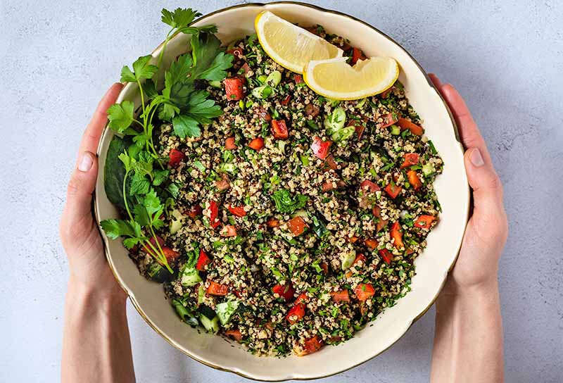 Overhead shot of two hands holding a large beige ceramic bowl of quinoa tabbouleh with tomatoes, green herbs, and yellow lemon wedges, on a white and gray speckled background.