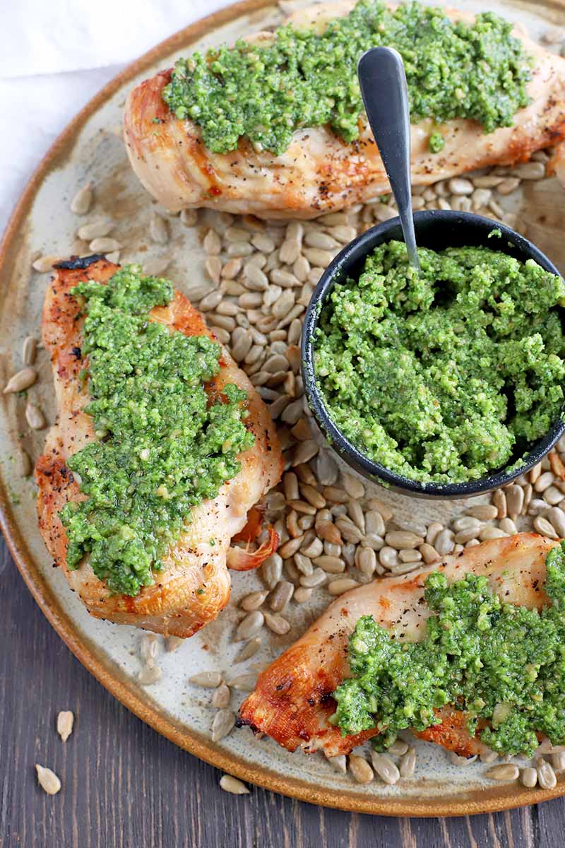 Overhead vertical image of grilled chicken breasts topped with pesto on a serving platter, with a bowl containing more of the dip at the center, on a brown wood surface with scattered sunflower seeds.