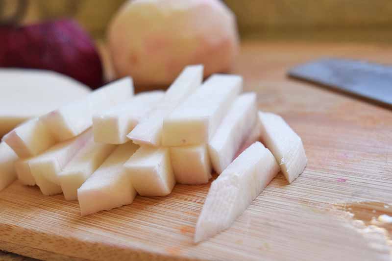Closeup image of peeled turnips sliced into thick matchsticks, on a wood cutting board with a knife and more peeled root vegetables in the background.