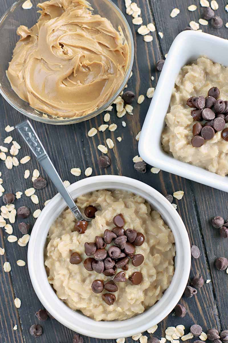 Overhead vertical shot of round and square white ceramic bowls of peanut butter with chocolate chips on top, with a spoon stuck into the one in the foreground, and a glass bowl of peanut butter, on a dark brown surface with scattered uncooked oats and dark chocolate.
