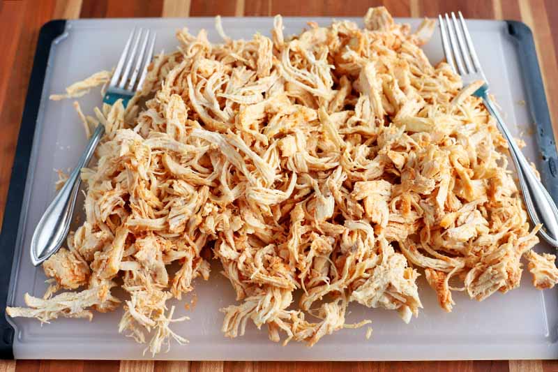 Shredded cooked chicken on a white plastic cutting board with two forks, on a wood surface.