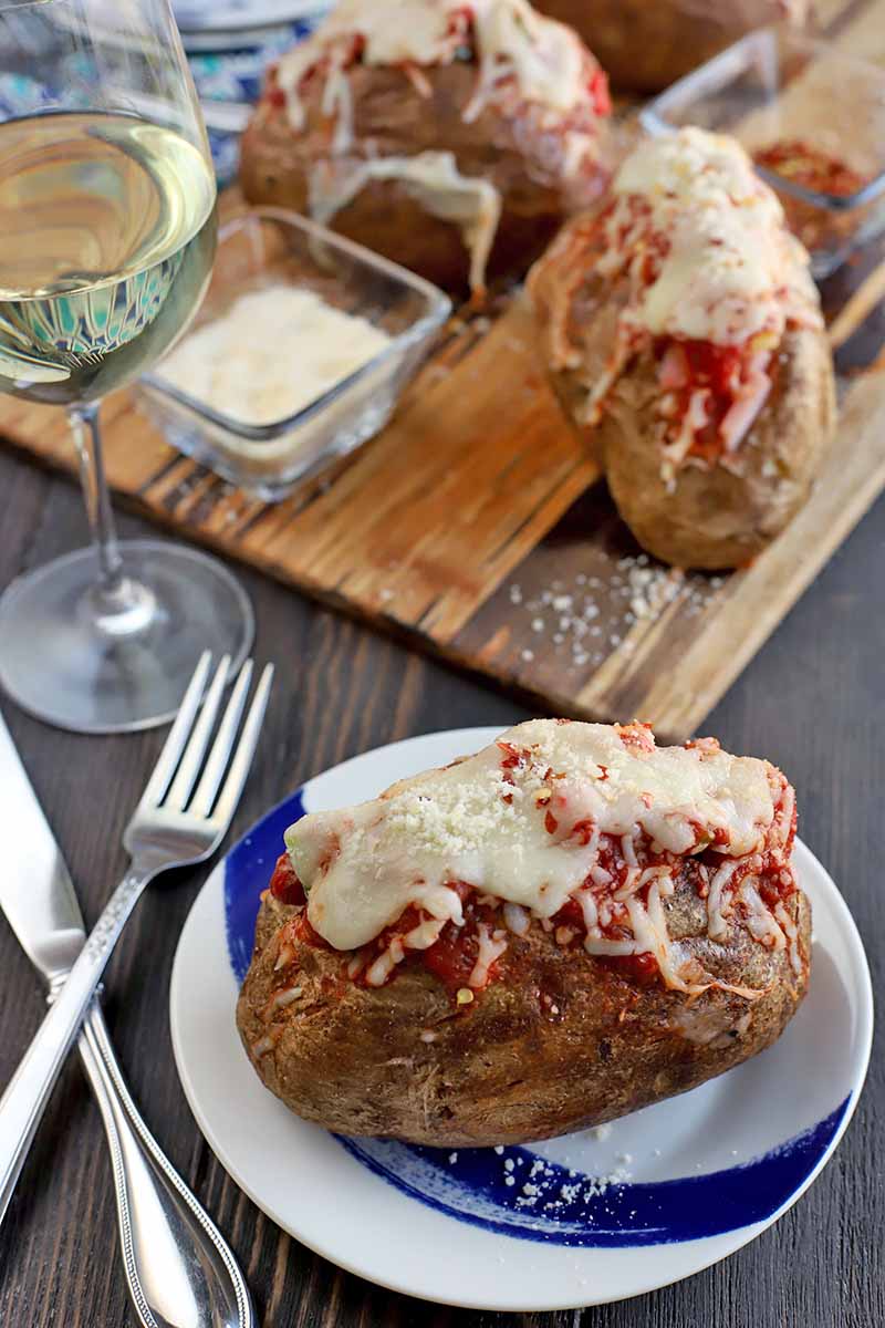 A baked potato stuffed with melted mozzarella and marinara sauce on a blue and white plate in the foreground with a crossed knife and fork to the left, with a glass of white wine in the background next to a serving board topped with smalls glass dishes of grated Parmesan and red pepper flakes, and more baked and filled spuds, on a dark brown wood table.