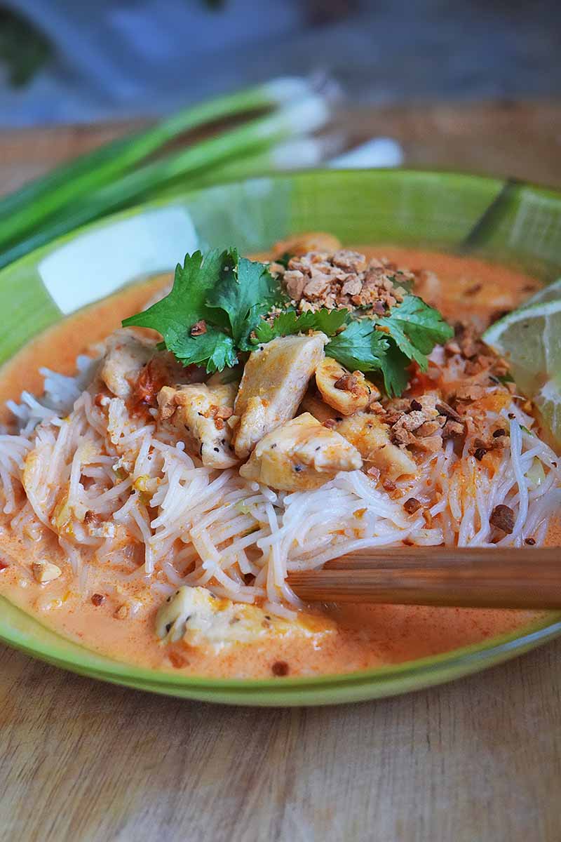Wooden chopsticks grab a bite of thin rice noodles in a red coconut curry broth with chicken, with a garnish of peanuts, cilantro, and lime, on an unfinished wood surface with scallions in soft focus in the background.