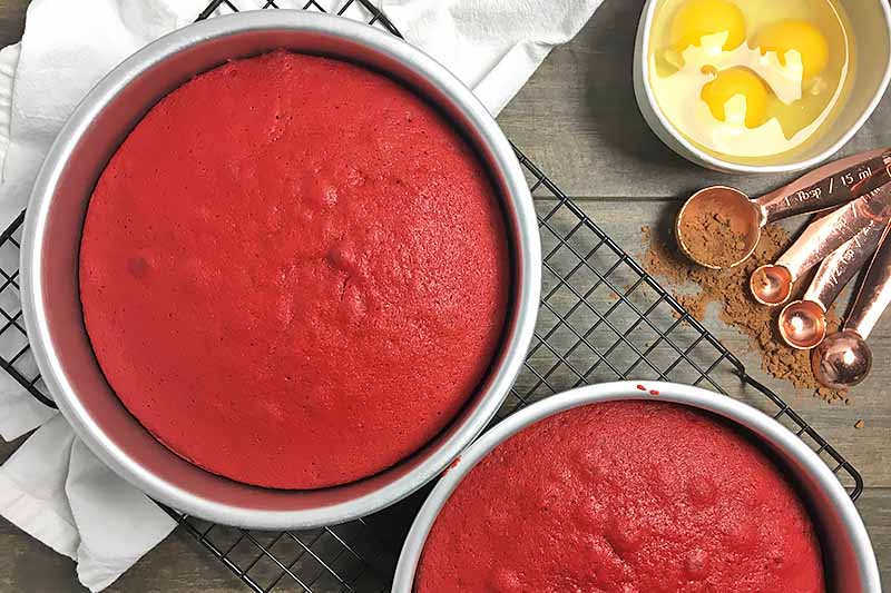 Horizontal image of baked red velvet cake in pans on a cooling rack next to eggs and cocoa powder.