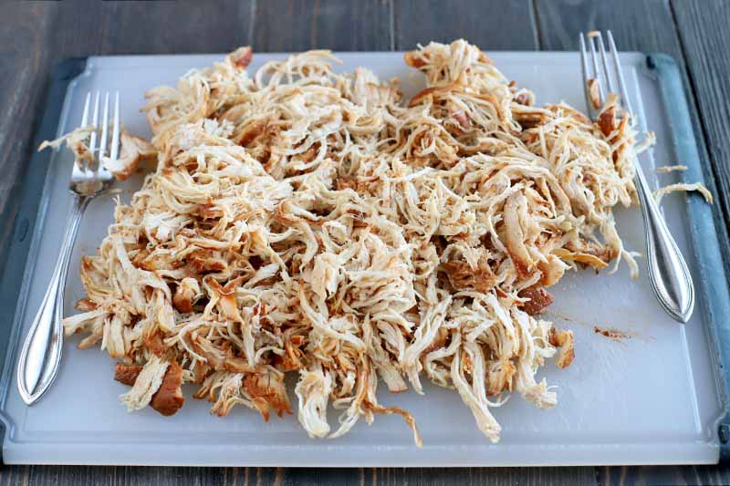 Shredded cooked chicken breast with barbecue sauce on a white and black plastic cutting board with two forks, on a dark brown wood surface.