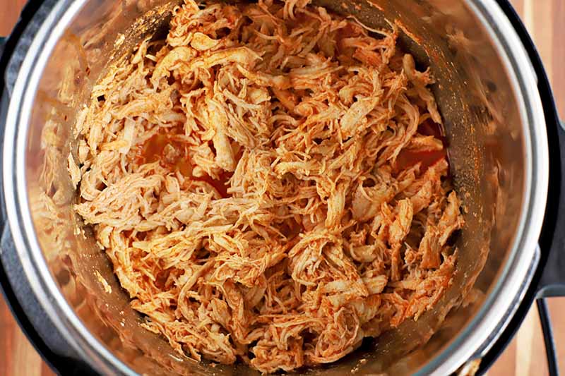 Shredded cooked chicken breast in the bottom of a metal slow cooker with red sauce, on a wood surface.