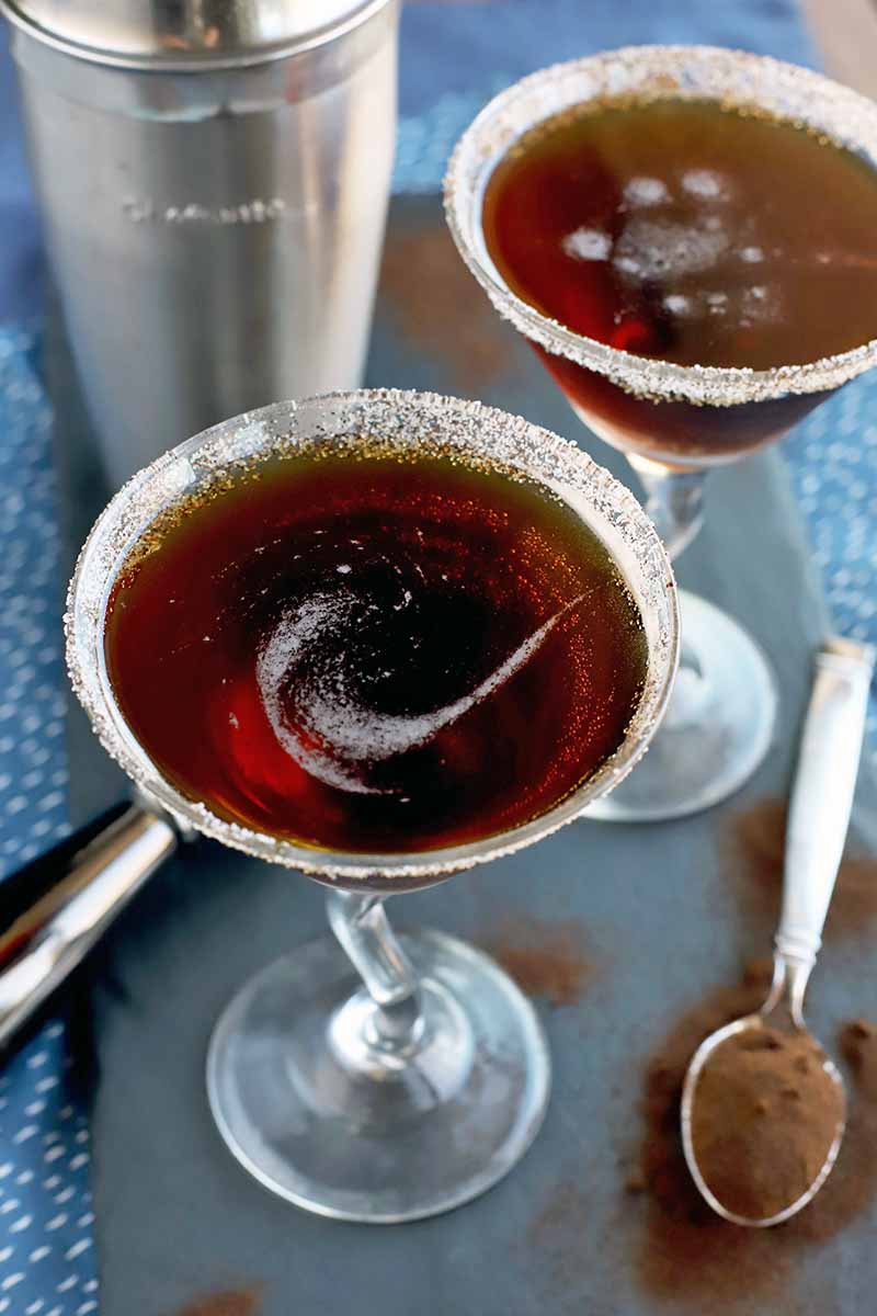 Two espresso martinis with a swirl of air bubbles on top and sugar rims, on a slate serving platter with a spoonful of coffee powder with more scattered around it, a stainless steel measuring jigger and a frosty cocktail shaker, on a blue and white cloth surface.