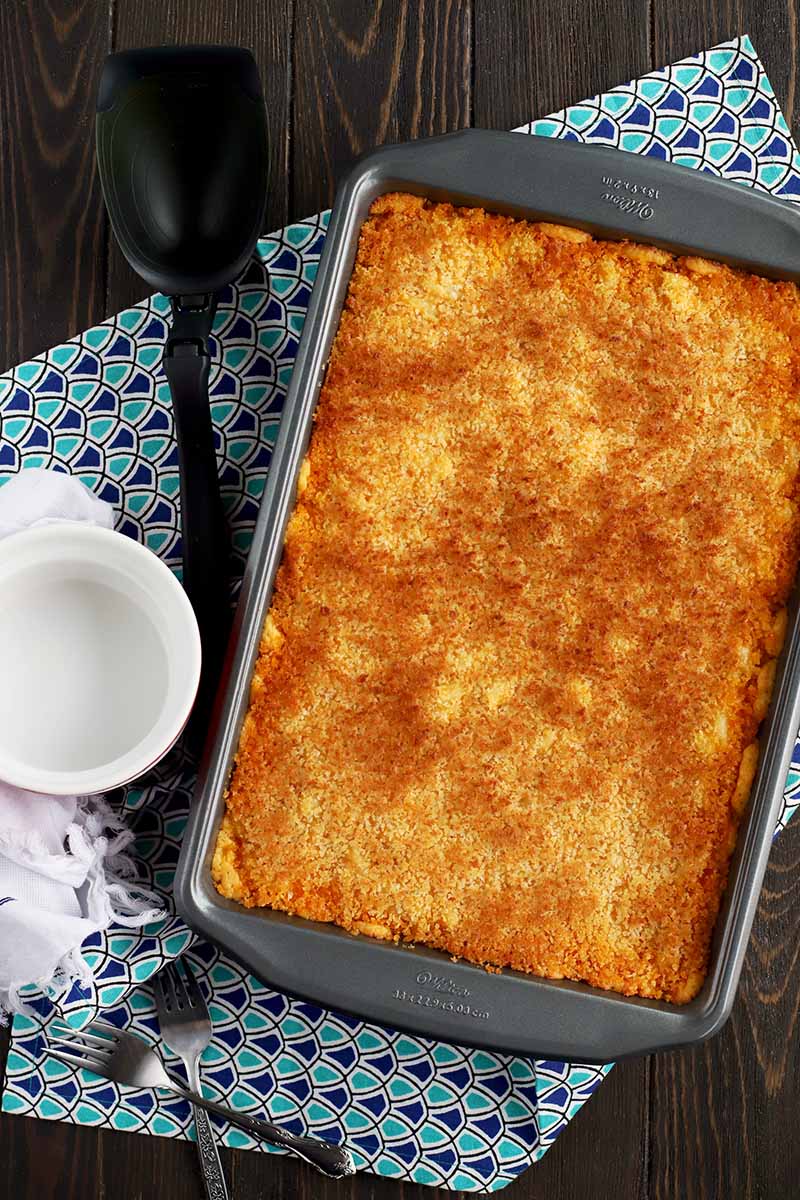 Overhead vertical shot of a metal rectangular baking pan filled with baked macaroni with toasted breadcrumbs on top, with a plastic serving utensil and a ceramic ramekin on top of a white dish towel and a folded dark and light blue patterned cloth, on a dark brown wood surface.