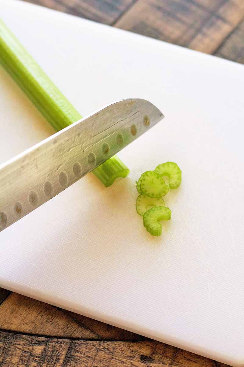 Vertical closely cropped shot of a knife chopping a stalk of celery on a white plastic cutting board, on a brown wood surface.