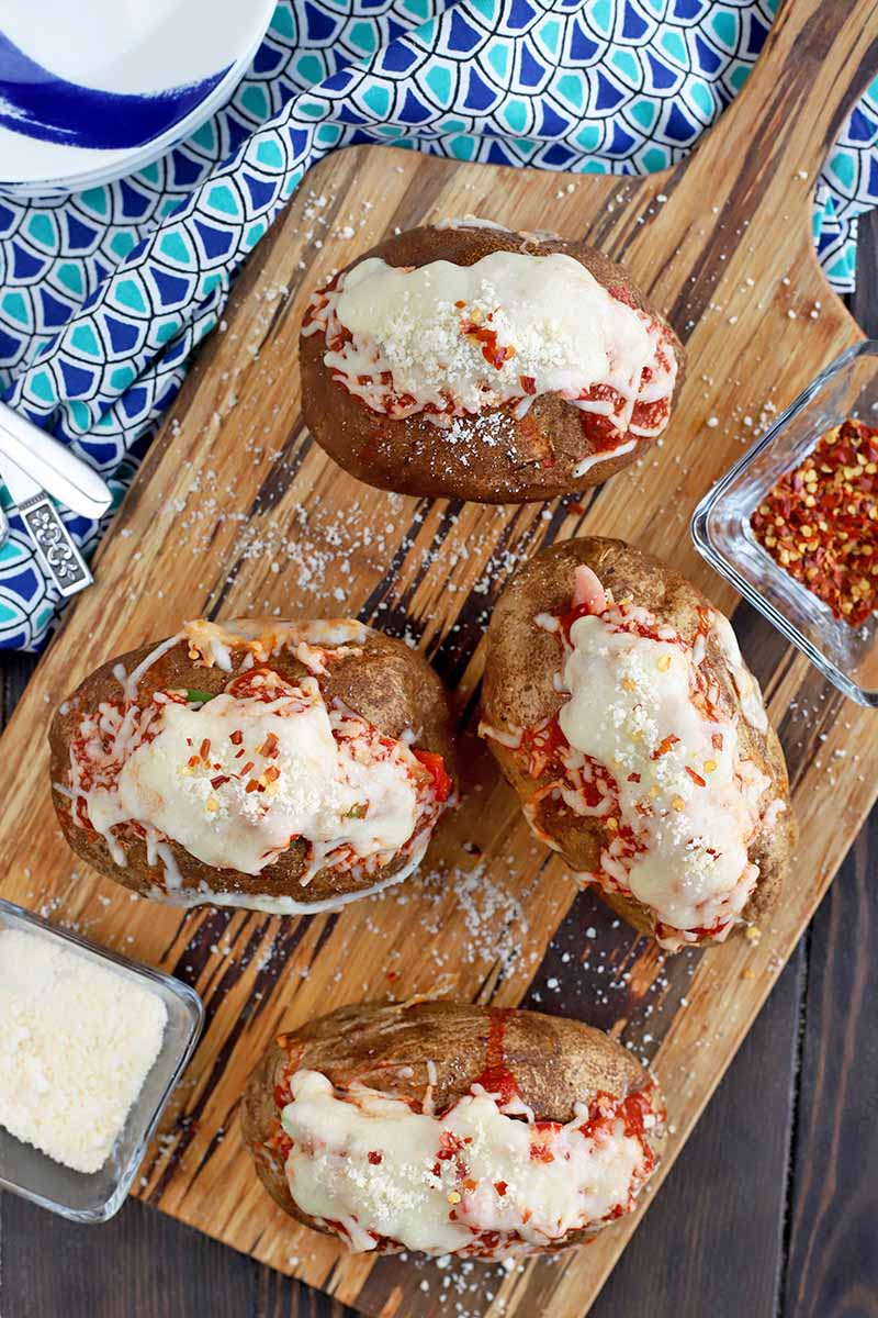 Overhead shot of four baked potatoes stuffed with marinara sauce and melted cheese, on a wooden board with a handle, with a small square dish of grated Parmesan to the left and an identical dish of red pepper flakes to the right, on a dark and light blue patterned cloth with a stack of blue and white plates to the left, on a dark brown wood surface.