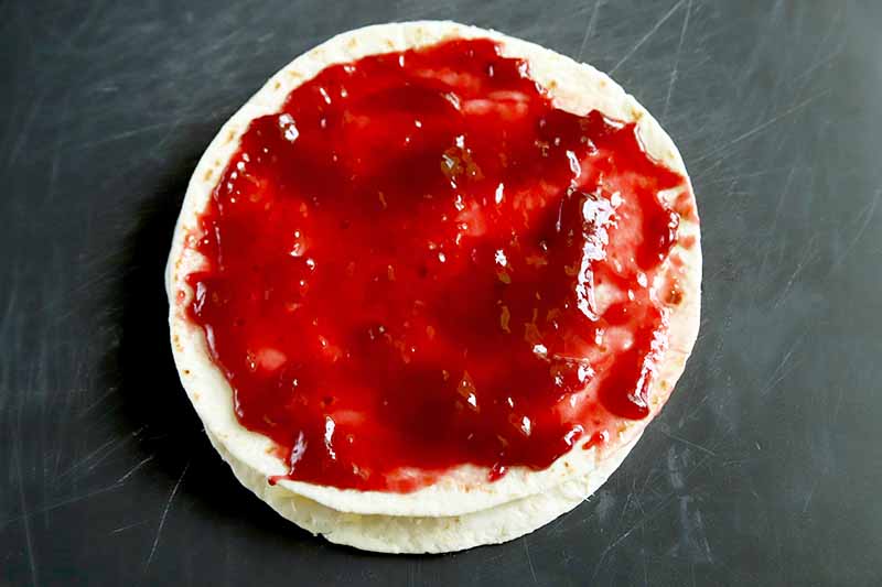 Horizontal image of two stacked four tortillas, with filling in between and raspberry jam spread in a thin layer on top, on a scratched gray surface.
