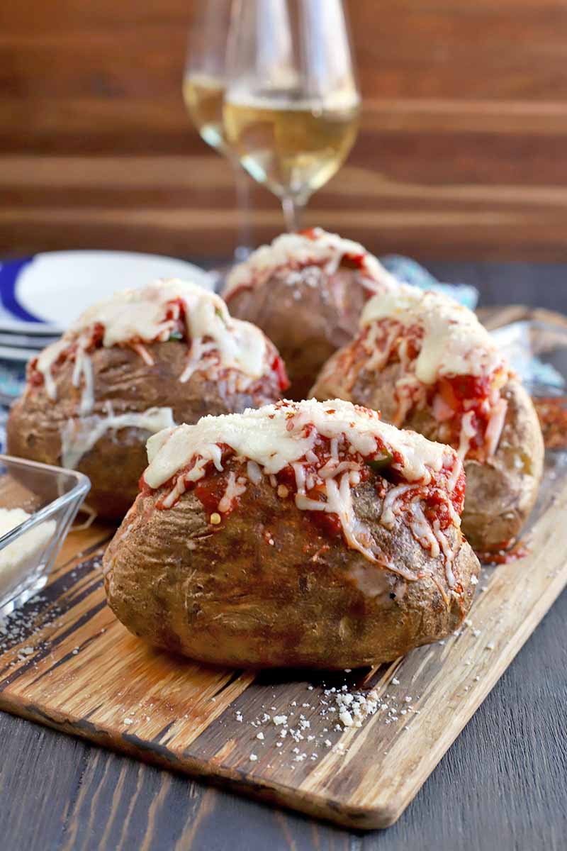 Vertical image of four baked potatoes with pizza topping fillings, on a wooden serving board with a small glass dish of grated Parmesan, and a stack of plates and two tall glasses of white wine in the background, with a wood backdrop and a dark brown wood surface.