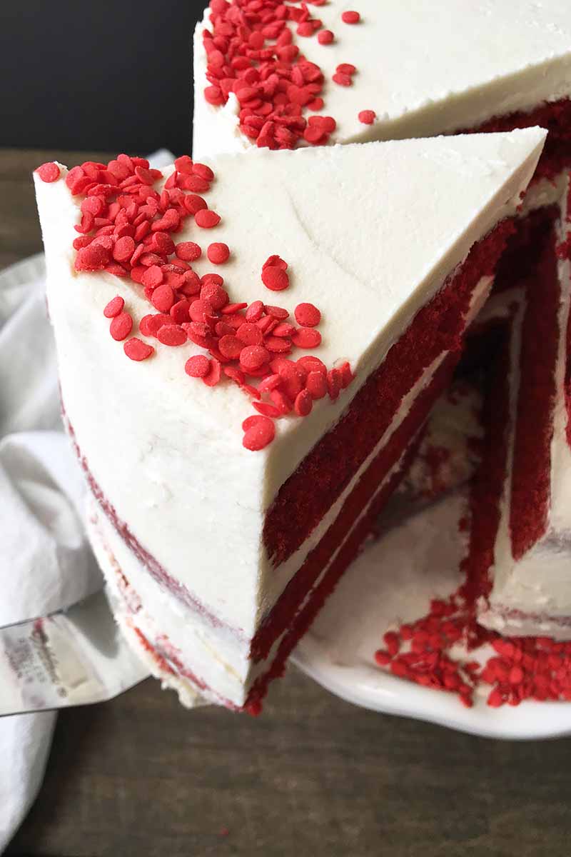 Vertical image of removing a slice of red velvet cake with white icing and sprinkles.
