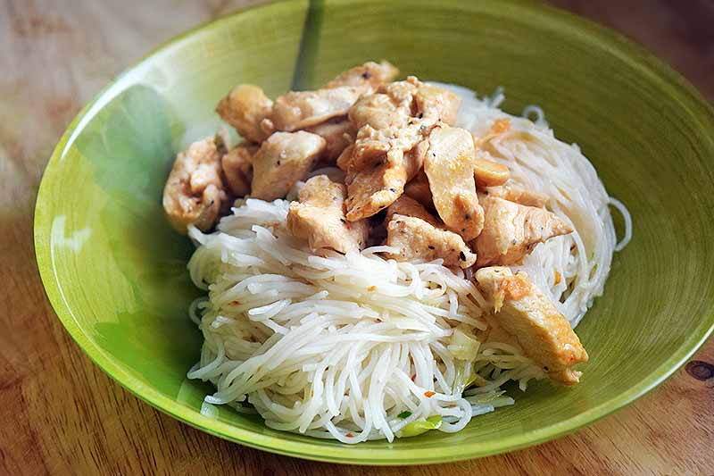 Rice noodles with cooked chicken in a green bowl, on a wood countertop.