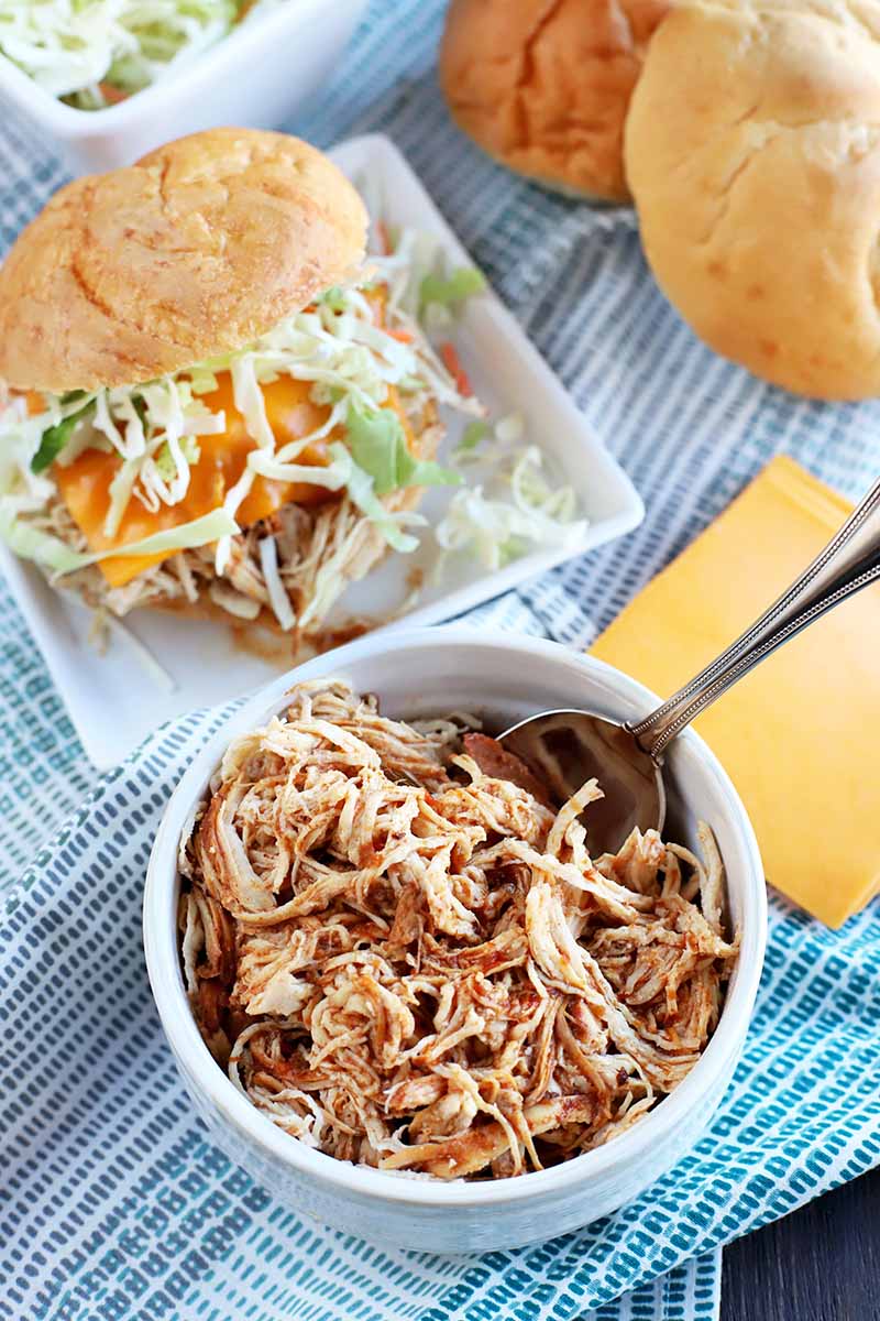 Overhead vertical shot of a white bowl of shredded chicken with barbecue sauce and a spoon, a square white plate with a sandwich topped with melted cheese and cabbage slaw, and a blue and white cloth topped with rolls, slices of cheddar, and a dish of shredded cabbage, on a dark brown wood surface.
