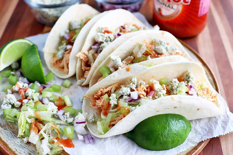 Four chicken tacos with hot sauce, blue cheese, lettuce, onion, celery, and lime, on a marble round serving platter with a bottle of hot sauce and small dishes of chopped vegetables, on a wood surface.