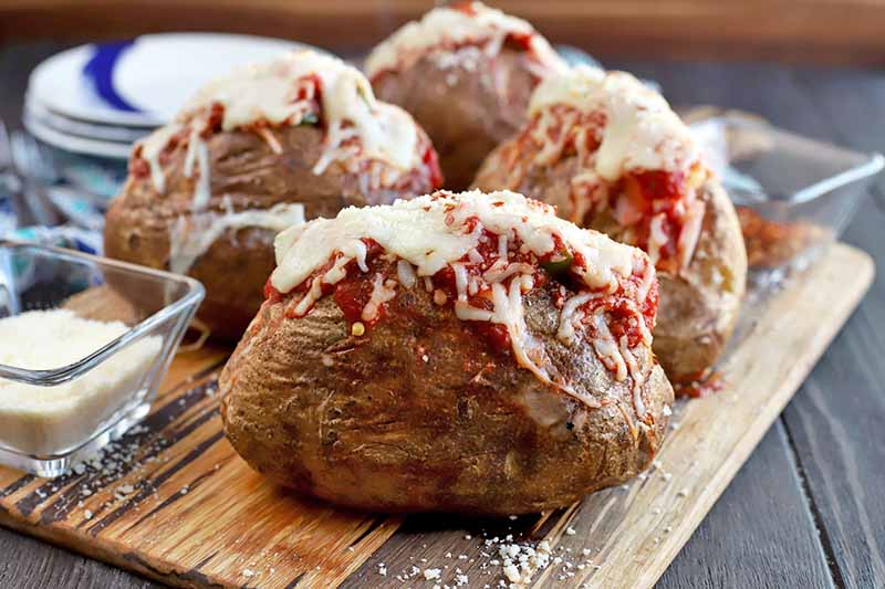 Four baked potatoes on a wooden board, filled with tomato sauce and melted cheese, with grated Parmesan sprinkled on top and in a small, square glass bowl to the left, with a stack of blue and white plate in the background, on a dark brown wood surface.