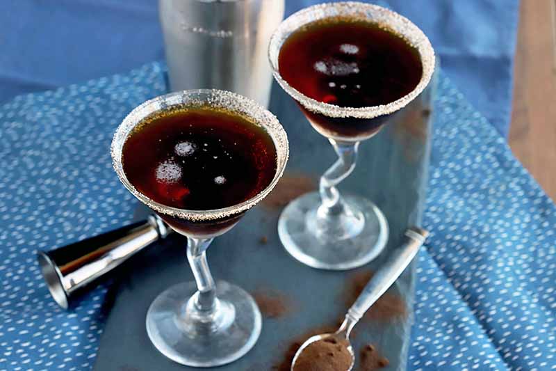 Two espresso martinis in glasses with decoratively bent stems, on a slate serving platter with a cocktail shaker and jigger, and a spoon filled with espresso powder with more scattered on the slate, on one plain blue cloth beneath a white and blue speckled patterned cloth, on a wood surface.