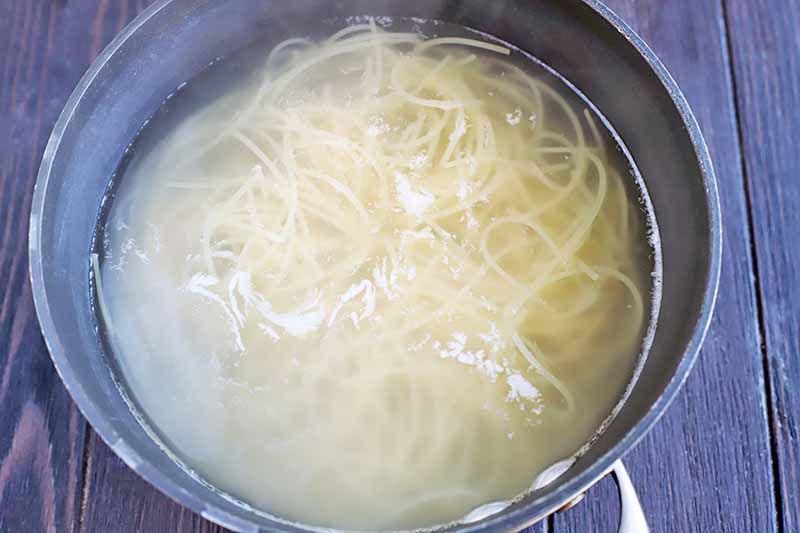 Cooked pasta noodles in water, in a large nonstick saucepan, on a dark brown wood surface.