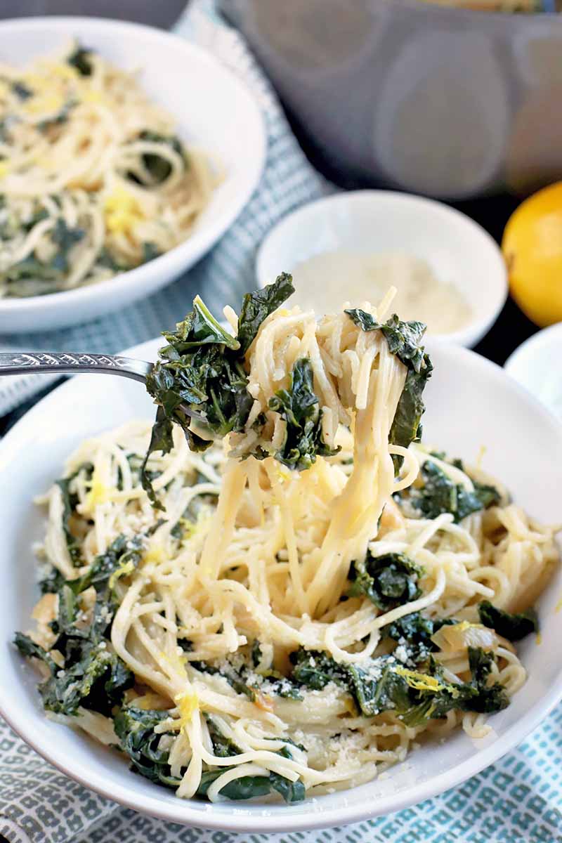 A forkful of pasta and greens is being held up to the camera, with more of the dish in a shallow bowl beside another identical dish, two smaller white bowls of grated cheese and citrus zest, a whole lemon, and a stainless steel serving dish, on a blue and white cloth.